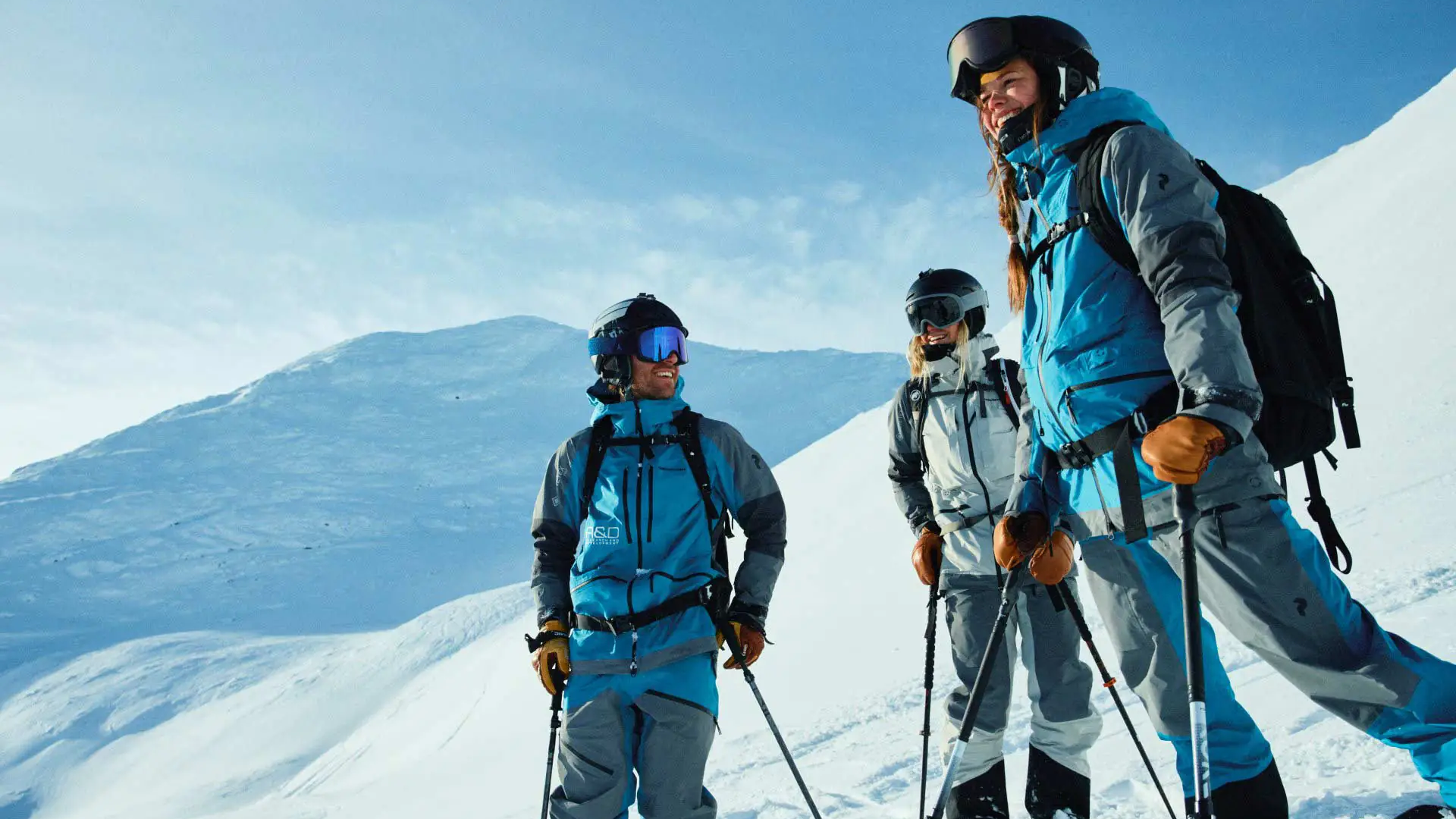 Drei Skitourengeher in Winterjacken stehen lachend auf einem verschneiten Berggrat